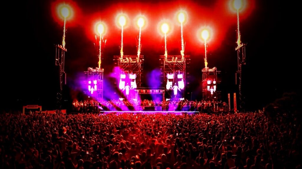 Panoramica notturna di palco e pubblico con luci rosse e viola ed effetti pirotecnici.